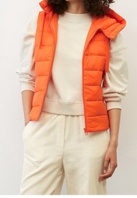 Gestepptes oranges Vest mit Kapuze, ausgestattet mit einem Frontreißverschluss und Seitentaschen, getragen über einem cremfarbenen Sweatshirt und gestreiften beigen Hosen.