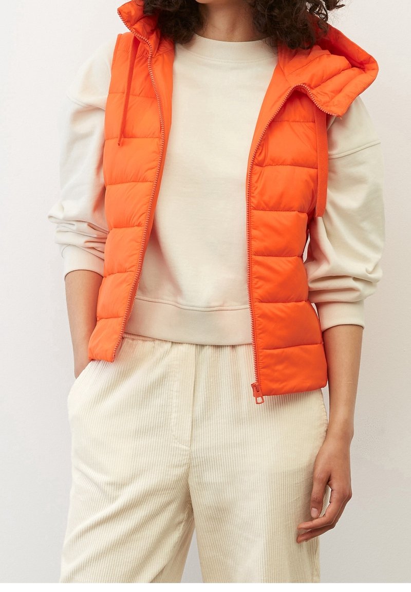 Gestepptes oranges Vest mit Kapuze, ausgestattet mit einem Frontreißverschluss und Seitentaschen, getragen über einem cremfarbenen Sweatshirt und gestreiften beigen Hosen.