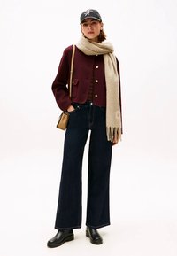 Ženska v temno rdečem krčenem jopiču, širokih temnih trapericah, bež rute, črnih škornjih, črni kapi in z bež torbo čez ramo.