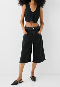 Top corto negro con escote en V profundo y botones frontales, combinado con culottes de mezclilla negros con bolsillos y un ajuste relajado.