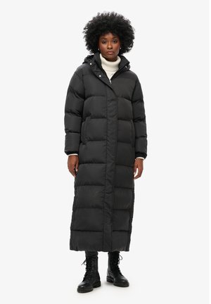 Kvinde stående iført en lang sort dunjakke med hætte, hvid rullekrave, sorte bukser og sorte snørestøvler.