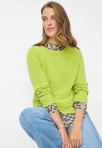Maglione verde brillante a coste con scollo rotondo, sovrapposto a una camicetta a fantasia. Il modello indossa jeans di denim azzurro chiaro e accessori.