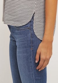 Blouse à motifs noir et blanc avec un ourlet arrondi, associée à un jean bleu. Le tissu est léger avec une texture lisse.