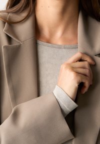 Woman wearing beige blazer and light gray sweater, hand adjusting the blazer near the collar.