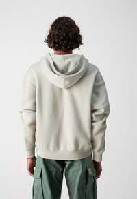 Felpa grigia chiara con cappuccio, finitura texturizzata, vestibilità ampia e polsini a coste, abbinata a pantaloni cargo verdi.