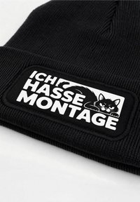 Black knitted beanie with a rectangular woven patch featuring white text "ICH HASSE MONTAGE" and an illustration of an angry black cat.