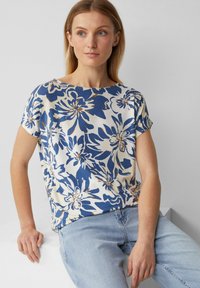 Blouse à motifs floraux crème, avec des fleurs bleues et brunes. Manches courtes et coupe décontractée, fabriquée en tissu léger et doux.