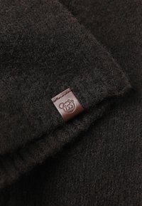 Ciemnobrązowa wełniana tkanina o teksturowanym wykończeniu z małą skórzaną metką z logo niedźwiedzia przyszytą do krawędzi.