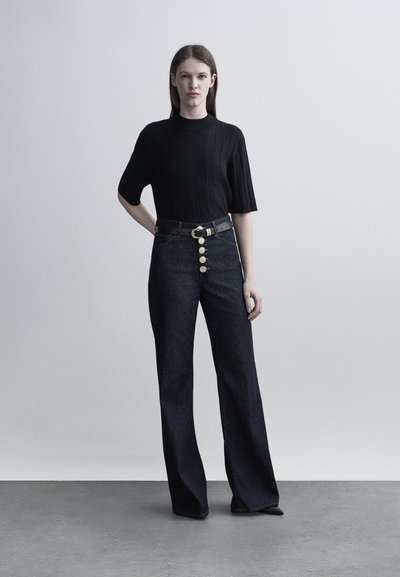 Γυναίκα με ίσια καστανά μαλλιά στέκεται φορώντας μαύρο κοντομάνικο κορυφαίο με ριμπ υφή, ψηλόμεσα σκούρα τζιν με χρυσαφένια κουμπιά και μαύρη ζώνη.