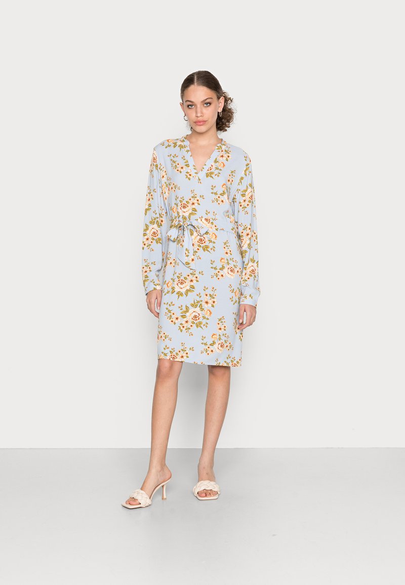 Vestido floral azul claro com mangas longas, decote em V, laço na cintura e comprimento até ao joelho. Apresenta padrões de flores laranja e verde.