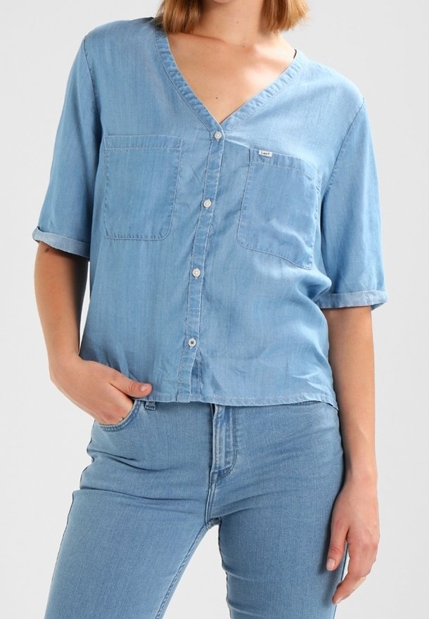 Chemise en denim bleu clair à manches courtes, décolleté en V, deux poches poitrine et fermeture à boutons. Texture douce, coupe légèrement ample.