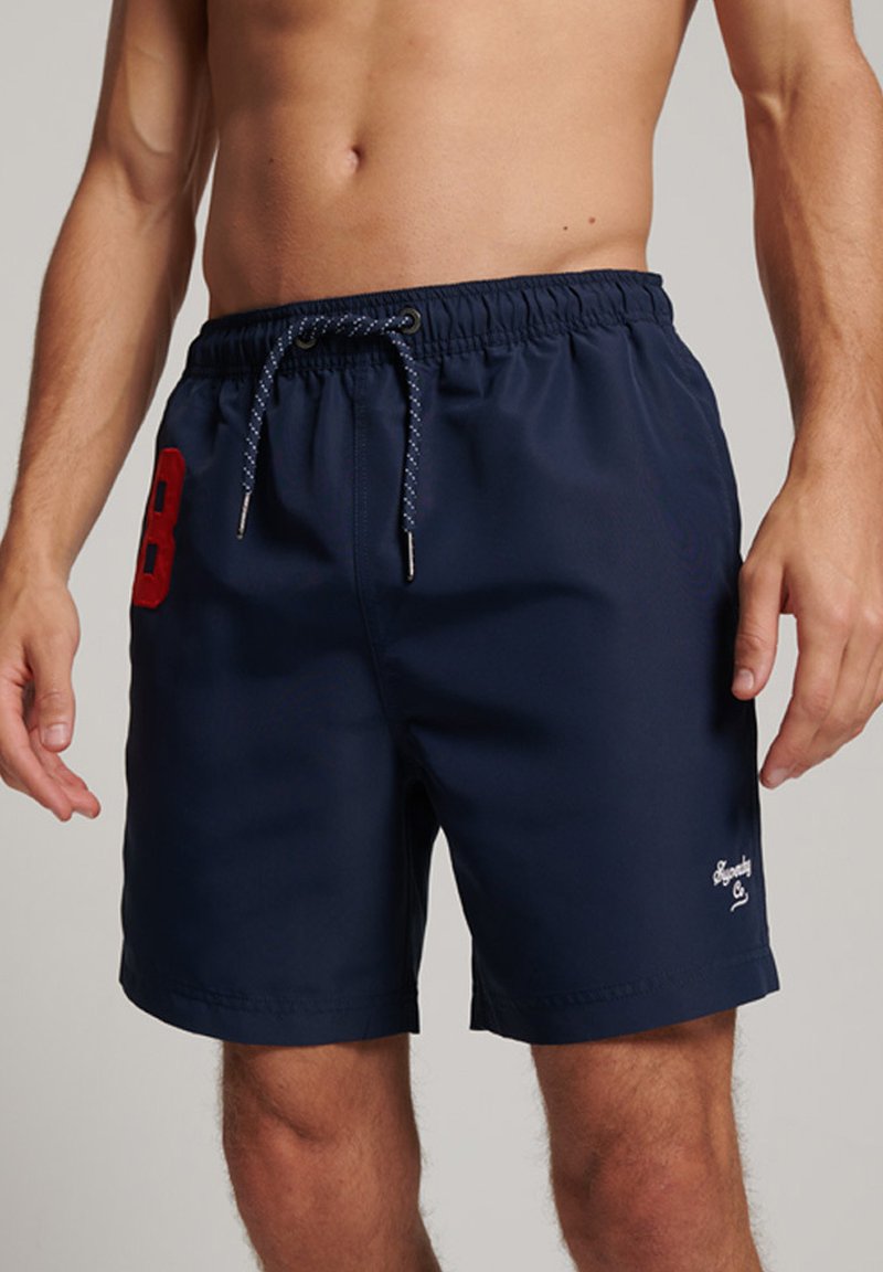 Pantaloni da bagno blu navy, vita elasticizzata con cordino, logo ricamato e un grande numero rosso 8 sul lato. Tessuto morbido e leggero.