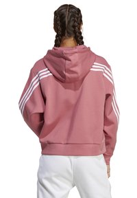 Personne aux cheveux tressés portant un sweat à capuche rose poudré avec des rayures blanches sur les manches et un pantalon blanc, vue de dos.