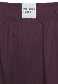 Bordeaux-housut, joissa on joustava vyötärö ja valkoinen etiketti, jossa lukee "COPENHAGEN STUDIOS". Ominaisuuksina sileä pinta ja sivutasku.