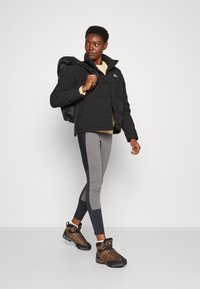 Czarna ocieplana kurtka z wysokim kołnierzem, szare legginsy z kolorowym blokowaniem oraz brązowe buty turystyczne. Modelka nosi czarny plecak.