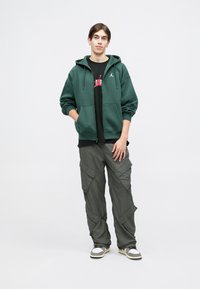 Felpa con cappuccio di colore verde scuro, dotata di tasche anteriori, indossata sopra una maglietta nera con grafica, abbinata a pantaloni cargo grigi chiari e sneakers.