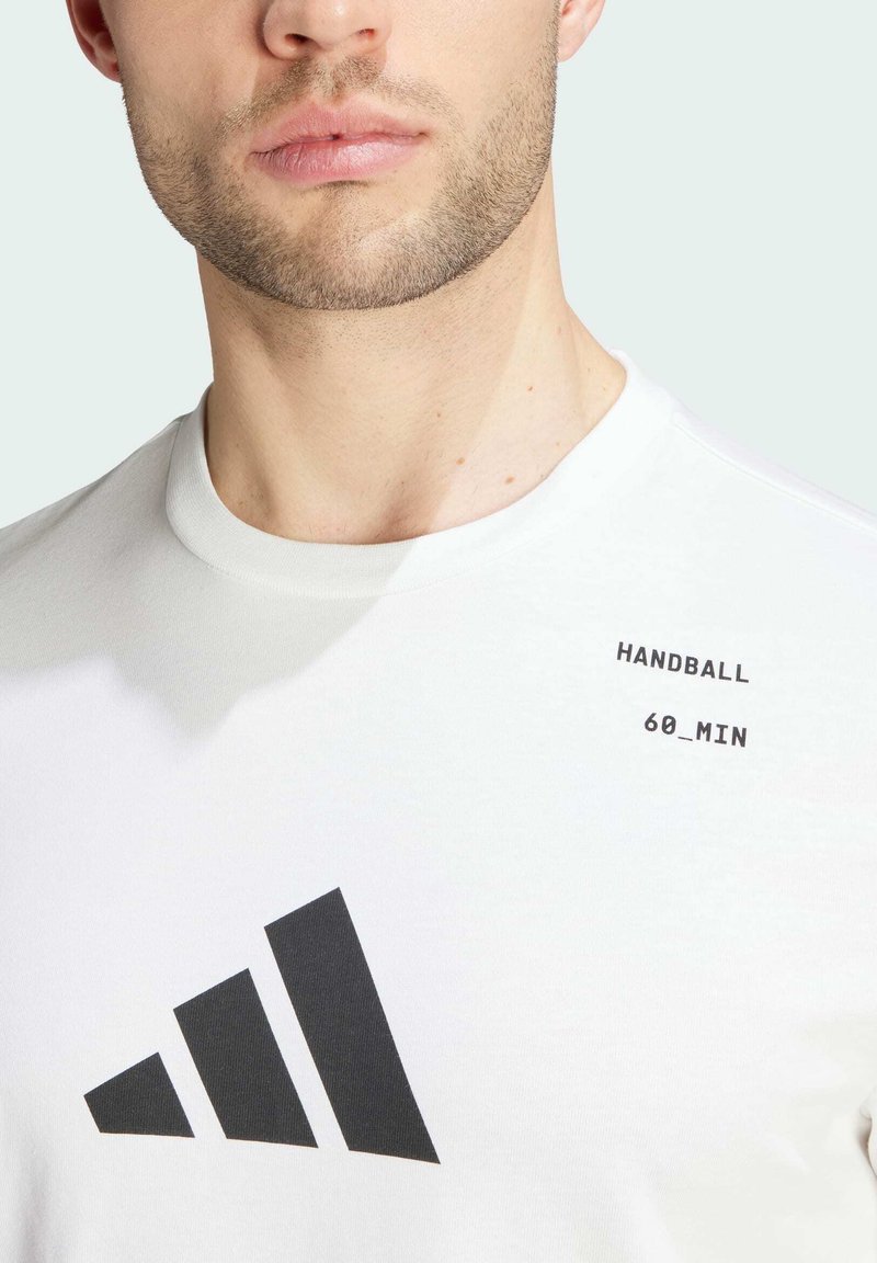 Camiseta blanca de cuello redondo, con un logo de tres franjas negras y el texto "HANDBALL 60_MIN" cerca del hombro. Material de algodón.