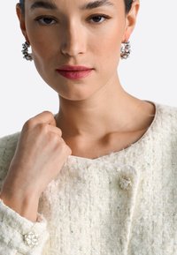 Balta faktūraota tweed jaka ar dekoratīvām pogām. Modelim sudraba riņķveida auskari. Tuvplāns uz seju un roku, kas satver audumu.