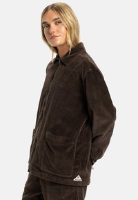 Brown corduroy jacket with a zippered front, collar, two front pockets, and a triangle logo accent near the hem. Textured fabric detail.