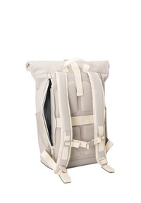 Sac à dos beige à fermeture enroulable, bretelles et panneau dorsal rembourrés, poche latérale zippée partiellement ouverte, présenté sur un fond blanc uni.