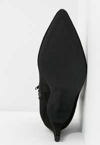 Botte noire à talon et bout pointu vue de la semelle, mettant en évidence son dessous texturé antidérapant et son talon fin sur fond blanc.