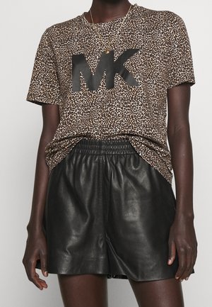 Frau trägt ein Leopardenmuster-T-Shirt mit großem schwarzem "MK"-Logo und schwarze Leder-Shorts, von Hals bis Mitte Oberschenkel gezeigt, neutraler Hintergrund.