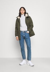 Olive rohelise värvi täispuhutav jakk karusnahast voodriga kapuutsiga, hall pikakarne särk, sinised teksad ja valged sneakerid. Omab tõmblukke ja nööbikammi detaile.