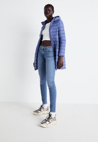 Gewatteerde, met een capuchon, lichtblauwe jas met lange mouwen, gecombineerd met nauwsluitende, medium-wash spijkerbroek en metallic sneakers, staande op een neutrale achtergrond.