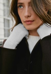 Közeli kép egy kézről, amelyet részben egy világosszürke kötött kesztyű takar, sötétbarna kabát közelében. A kabát alatt fehér pulóver látható.