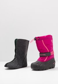 Botte en feutre noire avec un intérieur vert à gauche ; botte imperméable rose avec une sangle noire et une semelle en caoutchouc à droite.