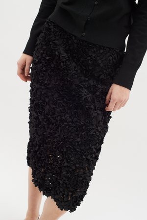 Black floral appliqué skirt featuring layered 3D flower details, knee-length cut, and a smooth texture. Worn with a black cardigan.