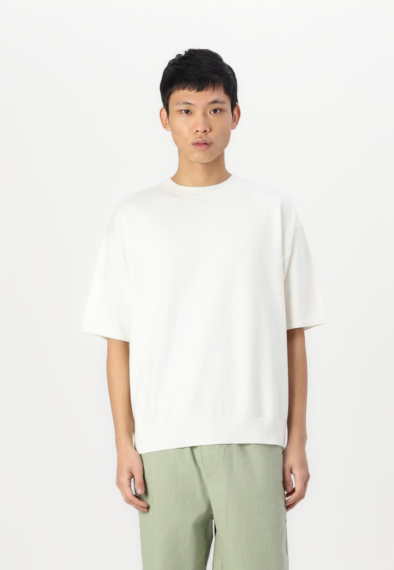 T-shirt tricoté blanc à manches courtes avec un col rond et une coupe décontractée, associé à un pantalon vert clair. Design simple, texture douce.