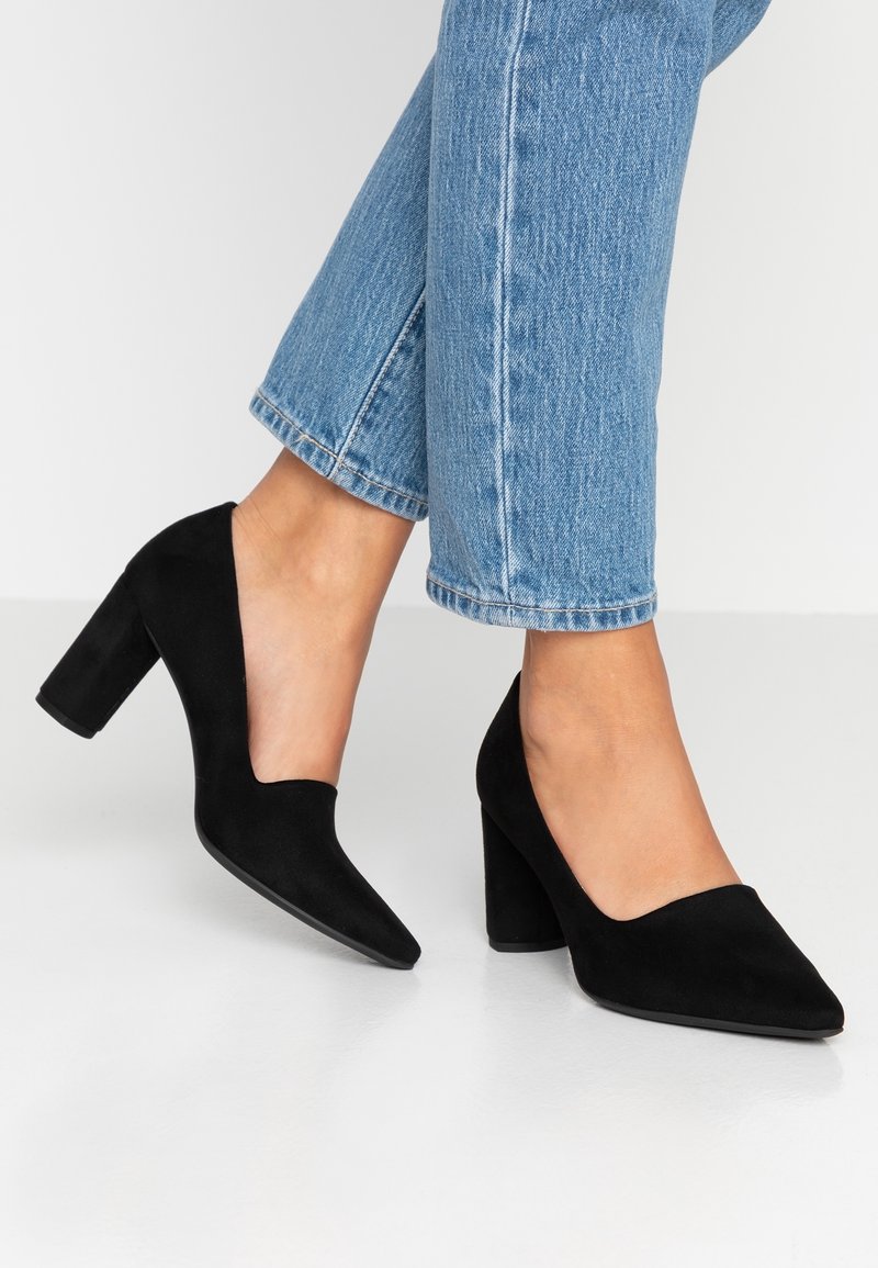Chaussures à talons hauts en daim noir avec un design en V et un talon carré épais, placées à côté d'un jean bleu clair coupé à la cheville.
