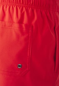 Red shorts made of lightweight fabric featuring an elastic waistband, side pocket, and a circular logo button with text.