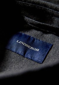 Denim kledinglabel in blauw satijn met witte tekst "LINDBERGH," bevestigd aan een grijs textuurstof.