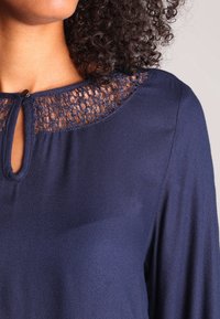 Blusa navy con scollatura a chiave e dettagli in pizzo, in tessuto morbido e con maniche ampie per una silhouette rilassata.
