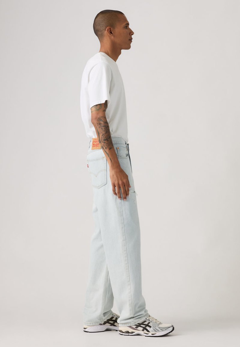 Hellblaue Jeans aus Denim mit entspanntem Schnitt, versehen mit einem aufgenähten Logo auf der Rückseite, getragen mit einem weißen T-Shirt mit kurzen Ärmeln und schwarzen Sneakers.