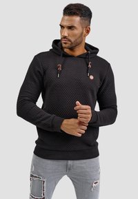 Zwarte hoodie met textuur, voorzien van koordjes en een ronde logo. Ribgebreide manchetten en onderkant, gedragen met versleten grijze jeans.