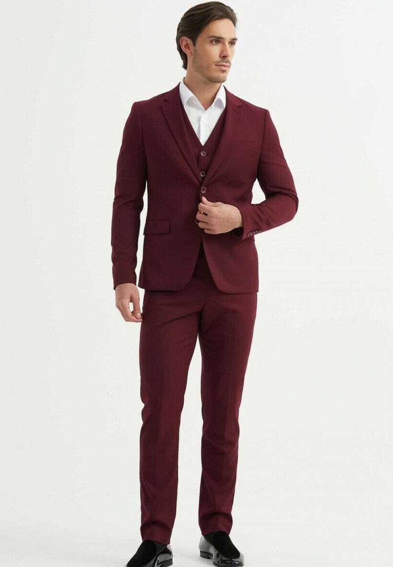 Costume bordeau avec une coupe ajustée, comprenant un blazer à simple boutonnage et un pantalon assorti. Chemise habillée blanche en dessous. Chaussures noires.