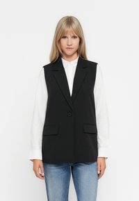Gilet sans manches noir avec une coupe ajustée, fermeture à un bouton et deux poches avant. Porté par-dessus une chemise blanche, avec une texture lisse.