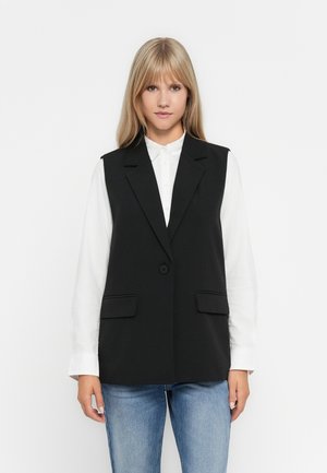 Gilet nero senza maniche con un design aderente, chiusura a pulsante singolo e due tasche frontali. Indossato sopra una camicia bianca, con una texture liscia.
