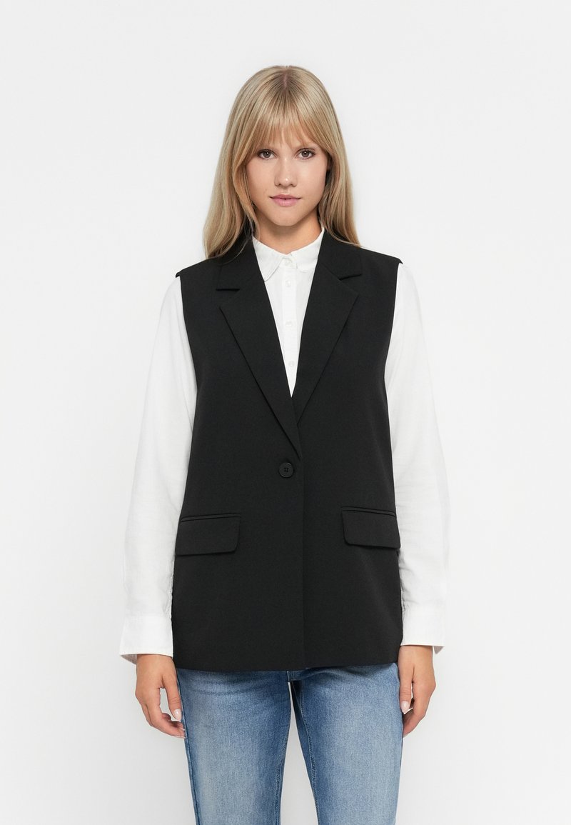 Gilet sans manches noir avec une coupe ajustée, fermeture à un bouton et deux poches avant. Porté par-dessus une chemise blanche, avec une texture lisse.
