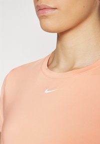 Nike lühikeste varrukatega särk pehmest virsiku värvi materjalist, ümmarguse kaelusega ja valge logo aktsendiga rinnal, sile tekstuur ja sobiv disain.