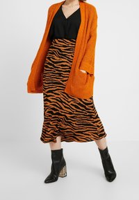 Kvinde iført en sort V-hals top, en midi-nederdel i orange og sort tigerstriber, en orange strikket cardigan og sorte ankelstøvler med mønstrede hæle.