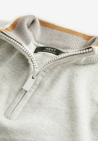 Grå ziptröja med ribbad krage, prydd med en gul-orange rand, texturerad tyg och en etikett som säger "NEXT."