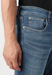 Light blue denim jeans featuring five pockets, yellow stitching, a small logo patch on the front, and a straight cut design.