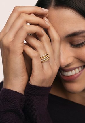 Conjunto de cuatro anillos de tono dorado con diseños retorcidos y lisos. Llevados en una mano de piel clara con uñas bien cuidadas.