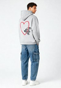 Felpa grigia con cappuccio caratterizzata da un grande cuore rosso e spille da balia nere ricamate sulla schiena. Abbinata a pantaloni cargo blu e sneakers bianche.