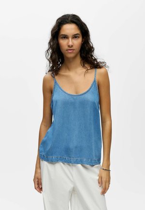 Jeune femme aux cheveux bouclés portant un haut bleu sans manches et un pantalon blanc, debout devant un fond clair uni.