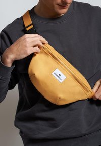 Borsa in tela gialla a vita con zip frontale, forma rettangolare, etichetta con marchio e tracolla regolabile. Texture liscia, adatta per l'uso quotidiano.